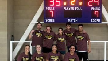 A group of eight players poses cheerfully in a gym after winning a basketball game.
