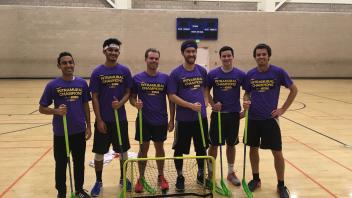 Six men in matching purple shirts pose with green sports sticks in a gym setting.