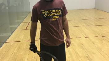 A man stands in a racquetball court holding a racquet, wearing athletic clothing and smiling.