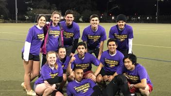 A group of eleven people wearing purple shirts posing together on a sports field at night.