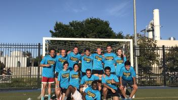 A diverse group of people in turquoise shirts posing happily in front of a soccer goal.