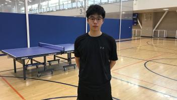 A young man stands in athletic attire in a gymnasium with a table tennis table nearby.