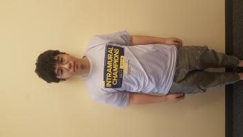 A person with tousled hair stands against a plain yellow wall, wearing a light purple t-shirt.