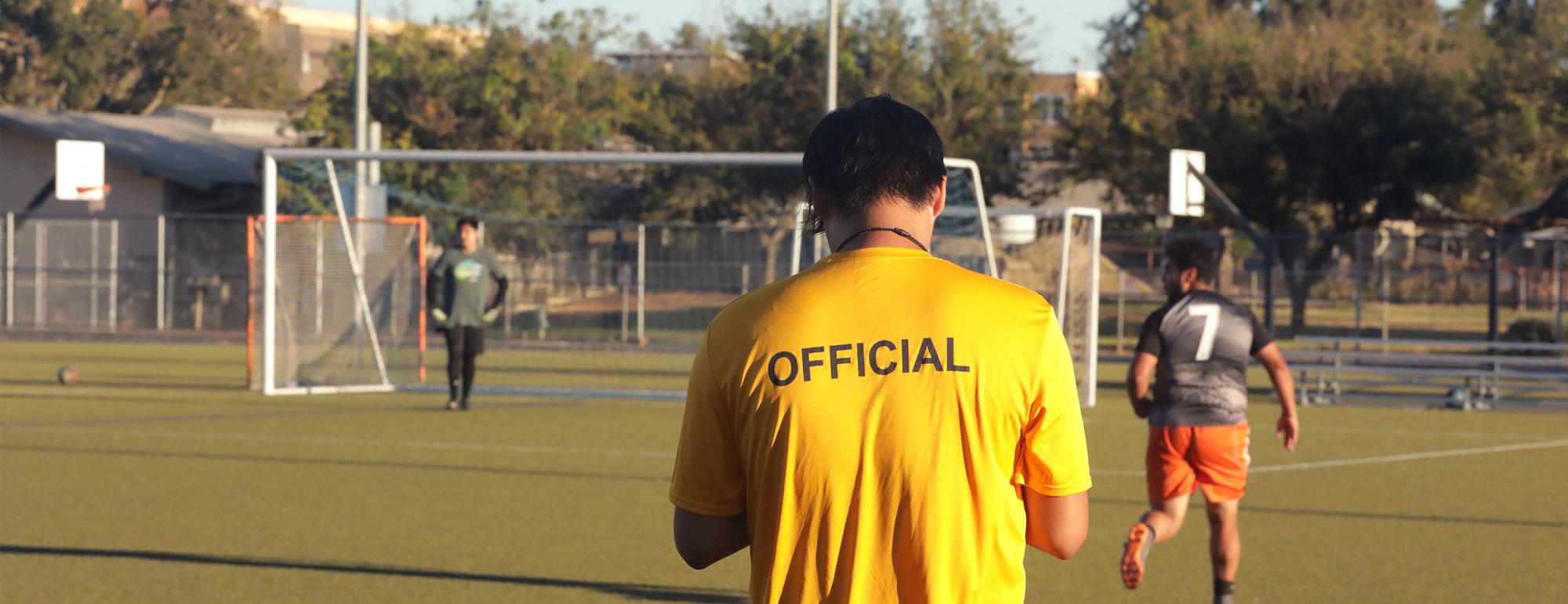 Official in yellow shirt stands with his back to camera on an outdoor soccer field
