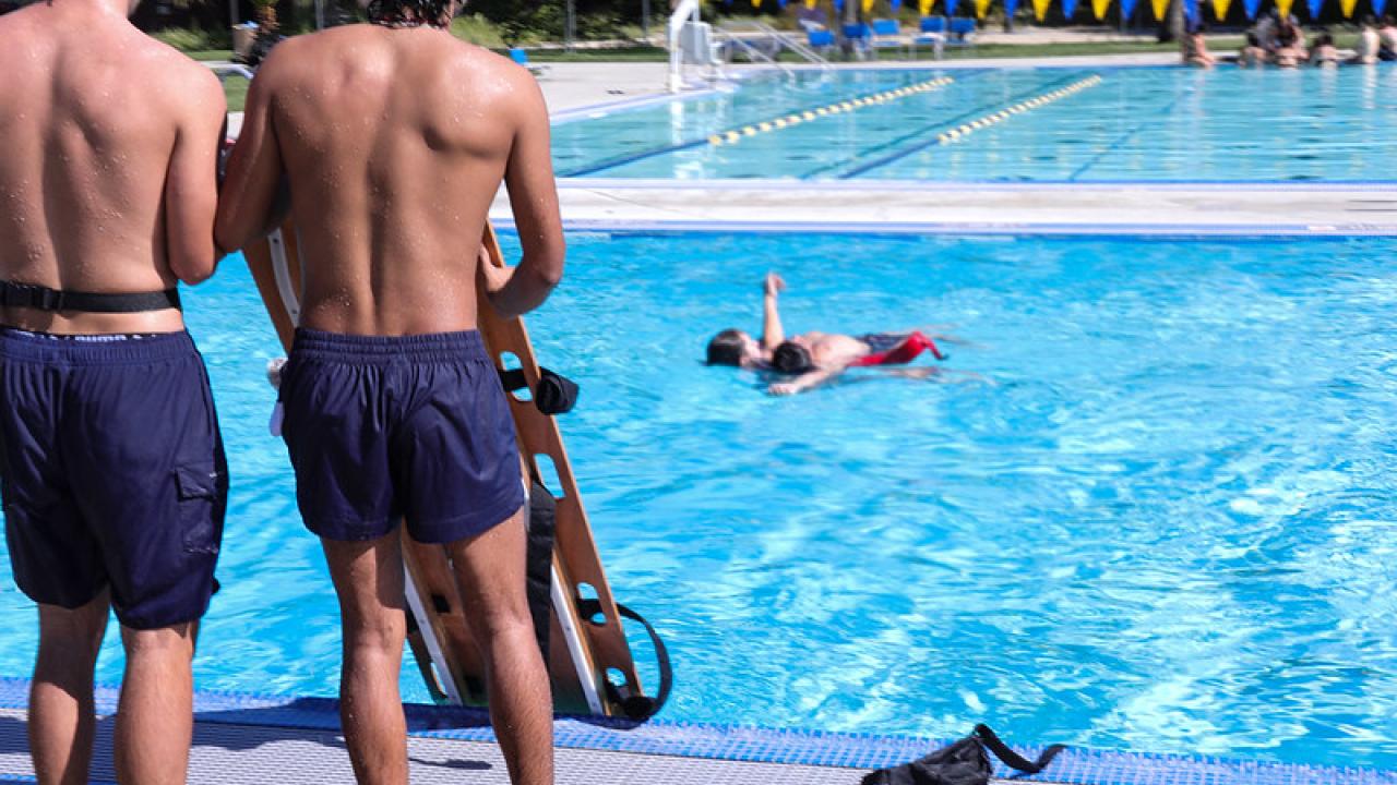 Two male student staff members are facing the pool, observing an individual in the pool as part of lifeguard training.