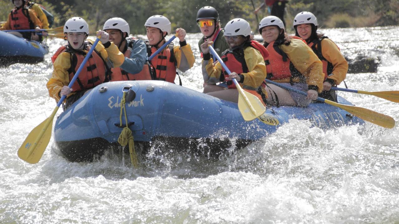6 students in a raft with paddles in their hands and big smiles as they go through a whitewater rapid