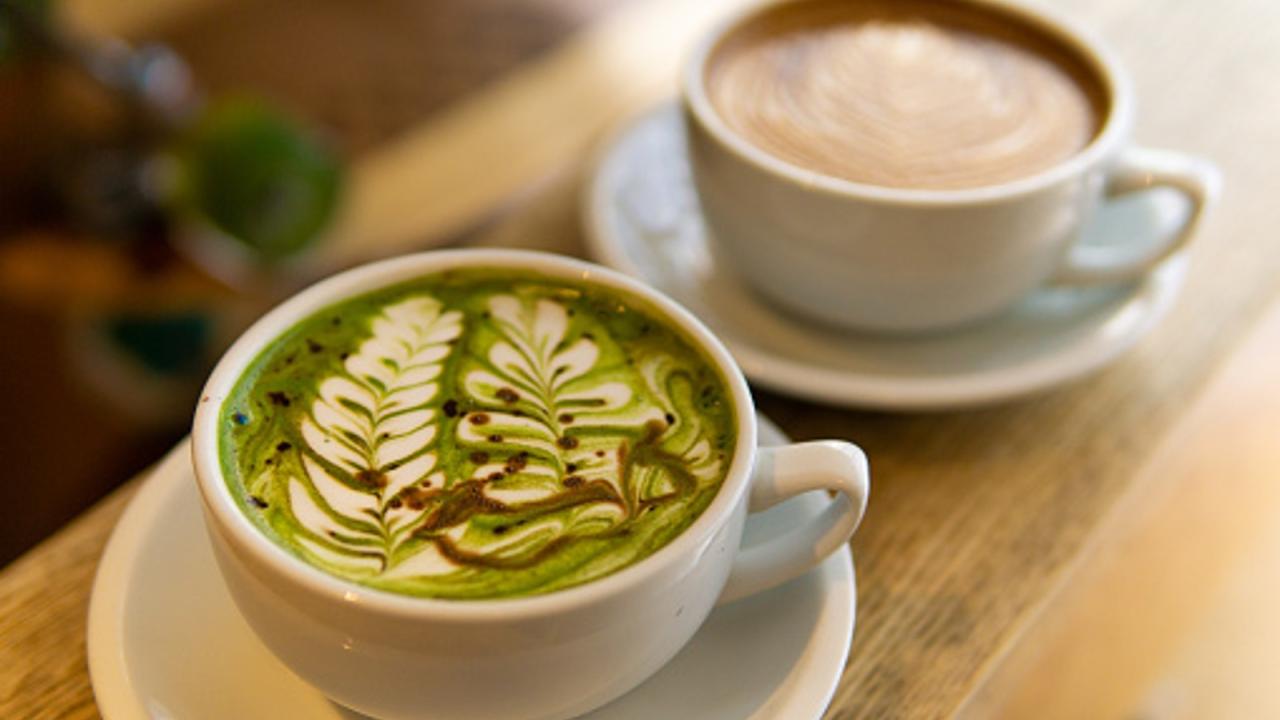 A close up of two mugs. The mug in the foreground is a green mug of matcha with a floral design in the milk. The mug in the background is a brown mug of coffee.