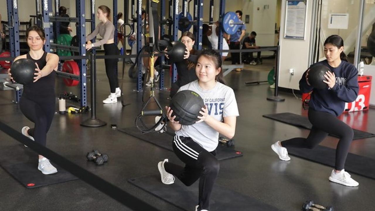 A group of students exercising in a group in the Rig in the ARC. The students are lunging with their right feet forward and are holding a medicine ball in their arms and twisting to the right.
