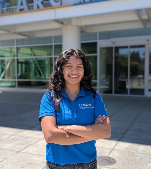 Smiling young woman in blue polo with arms crossed outside a campus building