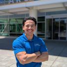 Smiling person in blue polo, arms crossed, standing outside a campus recreation building