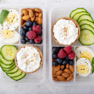 Two identical plastic tupperware with cucumbers and cottage cheese, almonds and berries, and hard boiled eggs in individual compartments.
