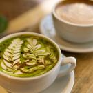 A close up of two mugs. The mug in the foreground is a green mug of matcha with a floral design in the milk. The mug in the background is a brown mug of coffee.